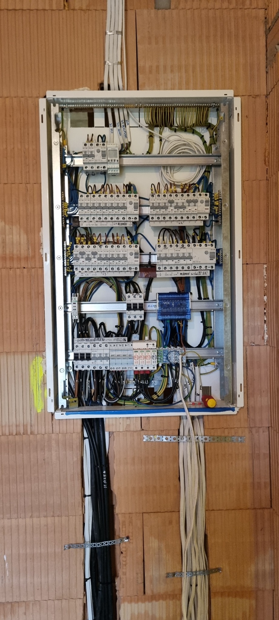Otwarta skrzynka rozdzielcza z licznymi przewodami, bezpiecznikami i elementami sterującymi zamontowana na ceglanej ścianie w Żyrardowie. Widoczne czarne i białe kable wchodzące i wychodzące ze...
