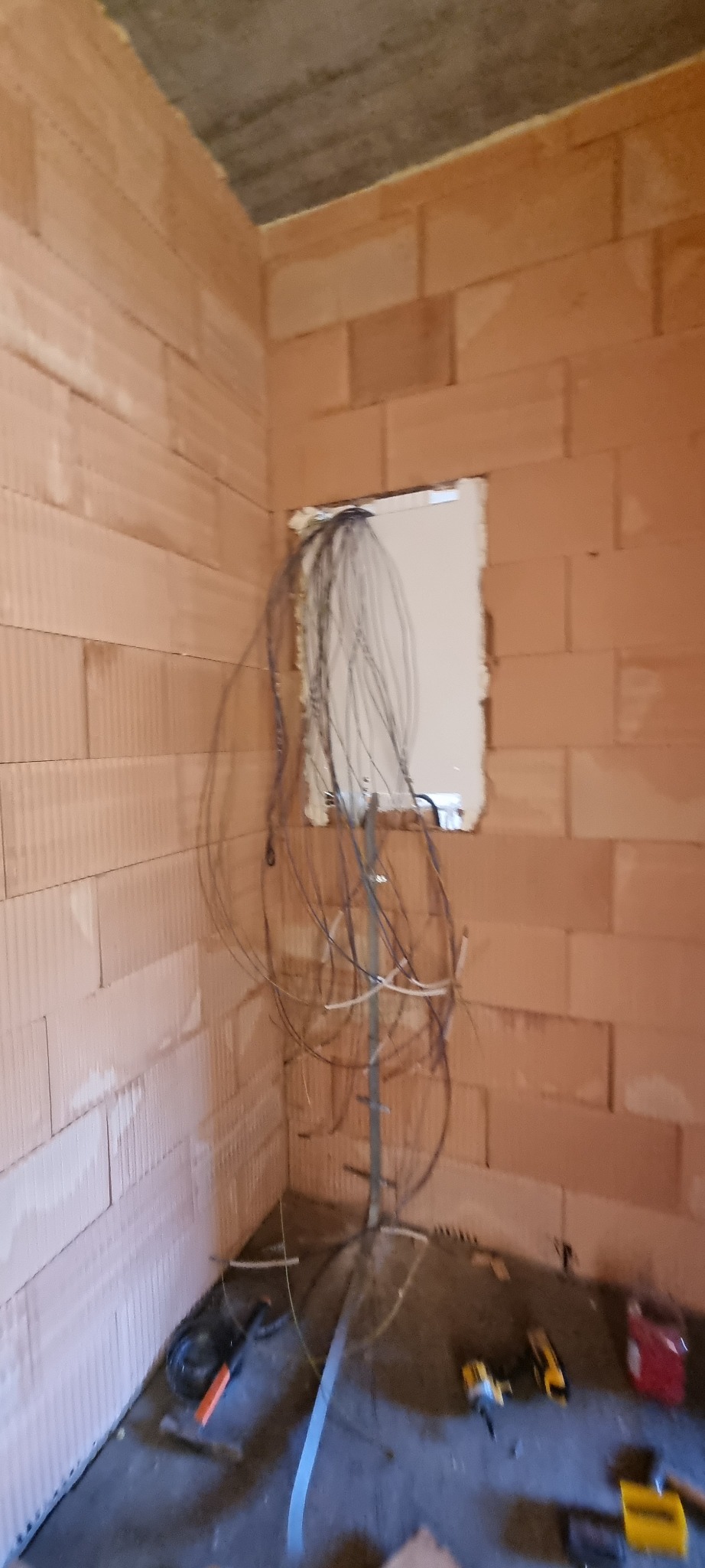 Wewnętrzna instalacja elektryczna w surowym budynku z czerwonej cegły: przewody wychodzące z otworu w ścianie, narzędzia elektryczne na podłodze, widok z dołu.