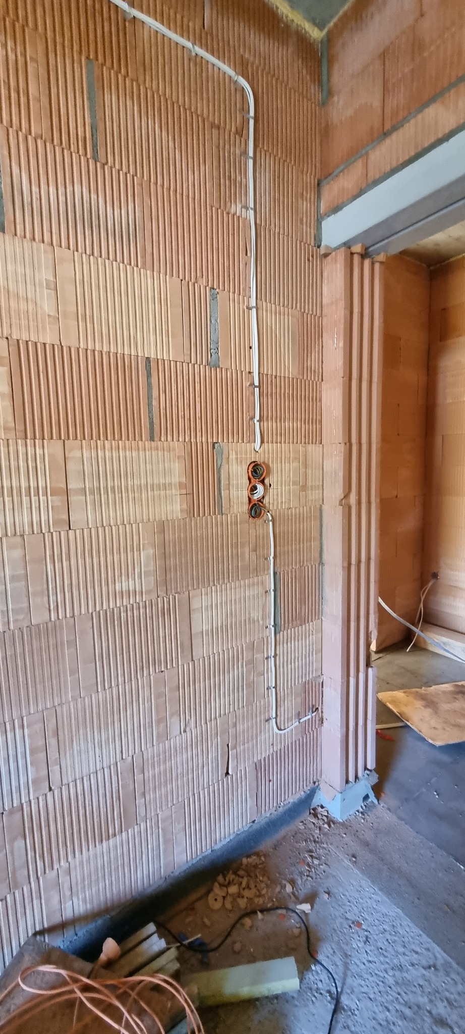 Ściana z czerwonej cegły z widoczną instalacją elektryczną: przewody w białej osłonie przymocowane do ściany i puszki instalacyjne w kolorze pomarańczowym.