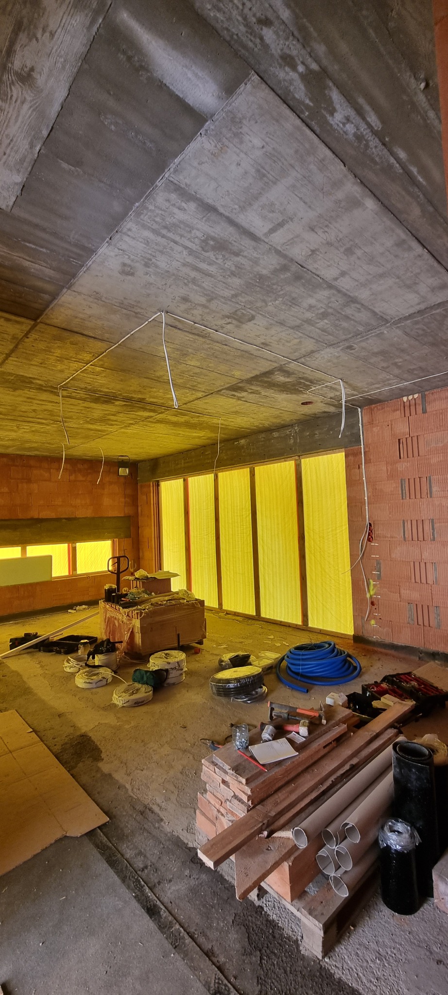 Surowy stan wnętrza budynku w trakcie prac, widoczne kable elektryczne pod sufitem, narzędzia i materiały budowlane rozłożone na podłodze, ściany z czerwonej cegły.