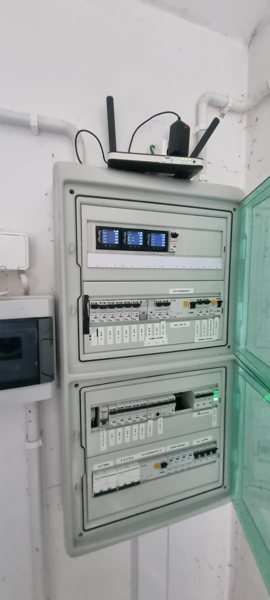 Otwarta skrzynka rozdzielcza z zainstalowanymi bezpiecznikami i modułami sterującymi, z routerem Wi-Fi umieszczonym na górze, na tle białej ściany.