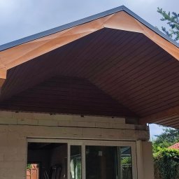 Firma budowlana Purgal - Drewniana podbitka dachu z widoczną konstrukcją więźby, szara blacha na krawędzi dachu, budynek w trakcie budowy z widocznymi oknami i rusztowaniem wewnątrz.