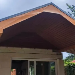 Drewniana podbitka dachu z widoczną konstrukcją więźby, szara blacha na krawędzi dachu, budynek w trakcie budowy z widocznymi oknami i rusztowaniem wewnątrz.