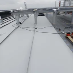 Montaż szarej blachy trapezowej na dachu przemysłowym, widoczne stalowe konstrukcje wsporcze, narzędzia na palecie i komin w tle.