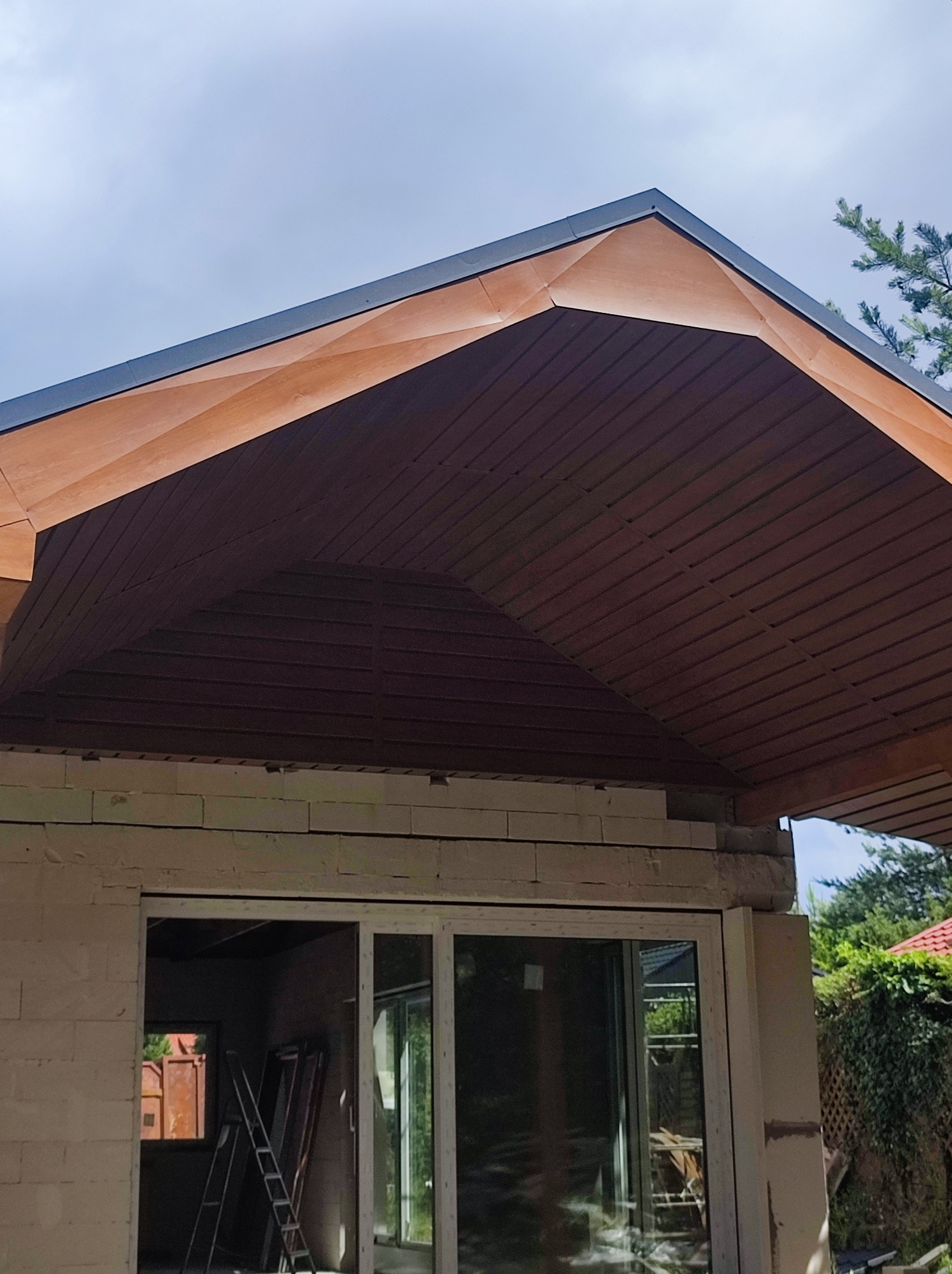 Drewniana podbitka dachu z widoczną konstrukcją więźby, szara blacha na krawędzi dachu, budynek w trakcie budowy z widocznymi oknami i rusztowaniem wewnątrz.