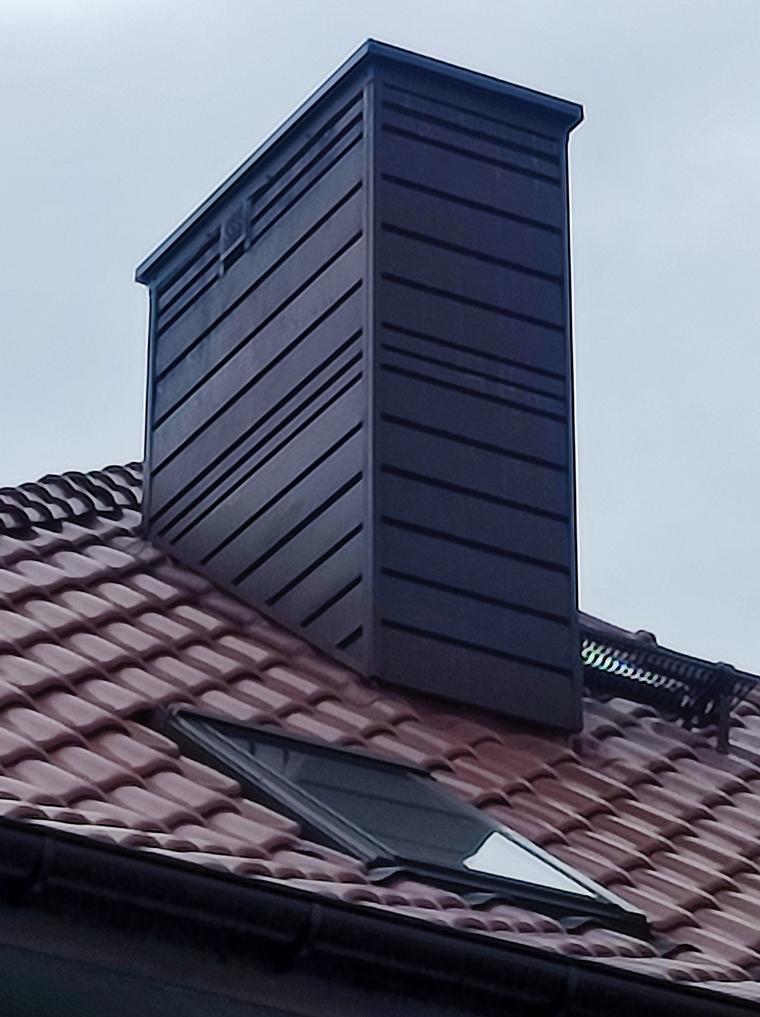 Obróbka komina z blachy w kolorze antracytu na dachu z dachówki ceramicznej, widoczne okno dachowe.