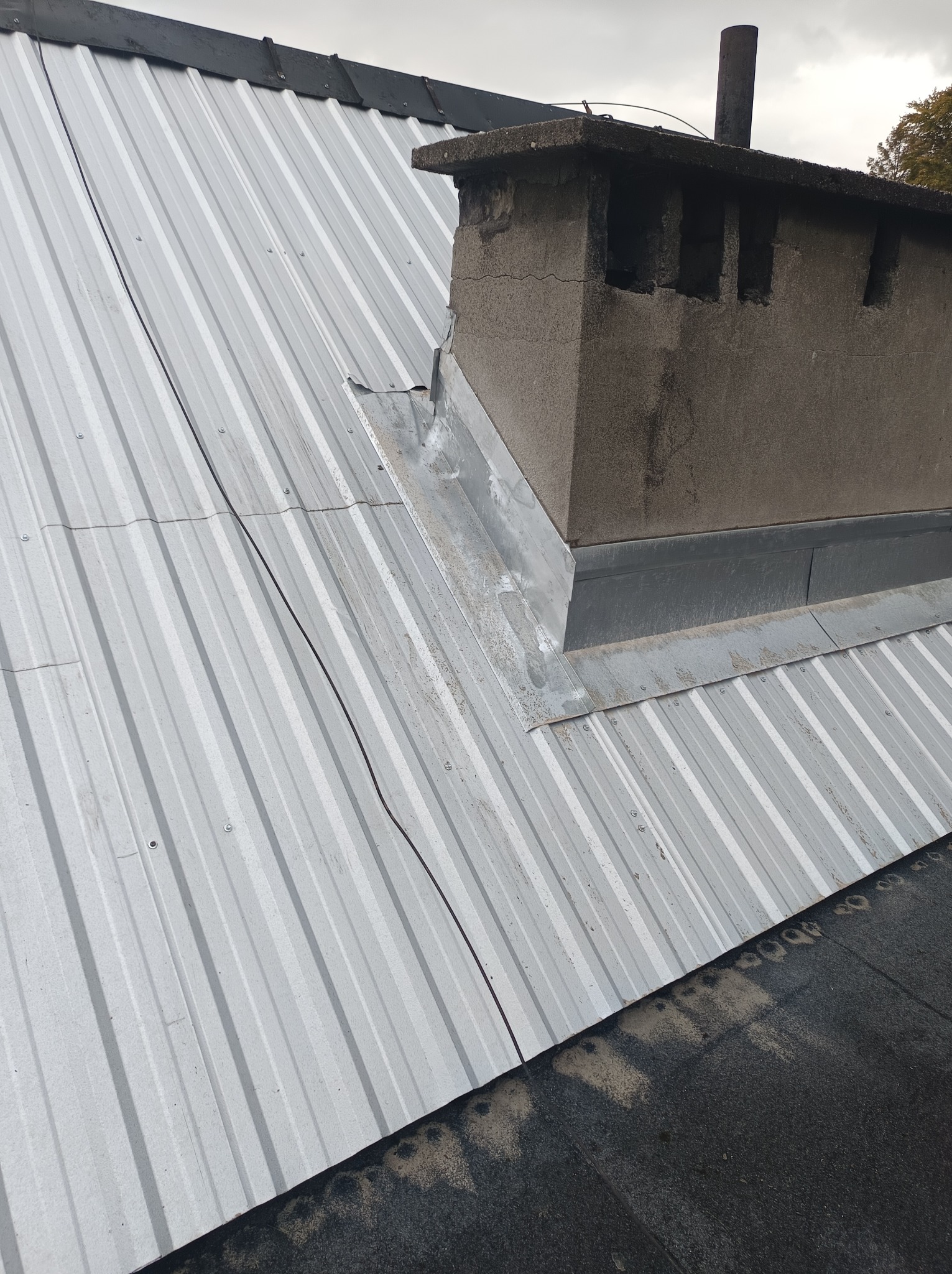 Szary dach z blachy trapezowej z kominem murowanym, obróbki blacharskie przy kominie, widoczny przewód na dachu.