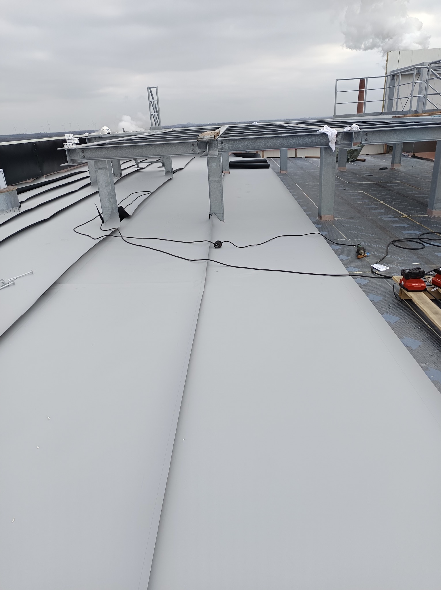 Montaż szarej blachy trapezowej na dachu przemysłowym, widoczne stalowe konstrukcje wsporcze, narzędzia na palecie i komin w tle.