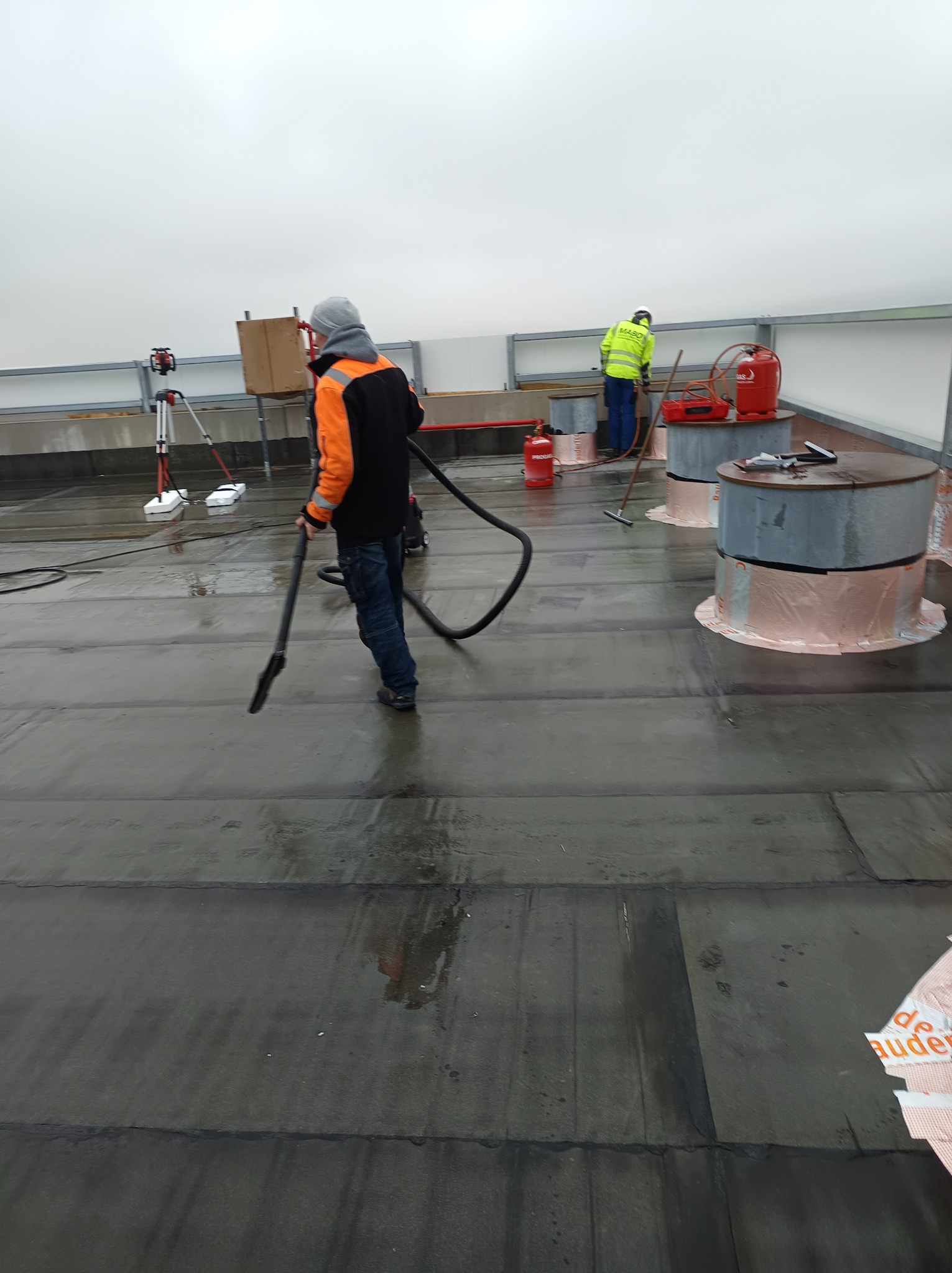 Pracownicy na płaskim dachu podczas czyszczenia powierzchni przed dalszymi pracami, widoczne narzędzia, butle z gazem i elementy konstrukcyjne.