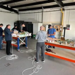 Grupa mężczyzn uczestniczy w praktycznym szkoleniu z zakresu instalacji elektrycznych, pracując przy stołach z elementami instalacji w przestronnym pomieszczeniu.