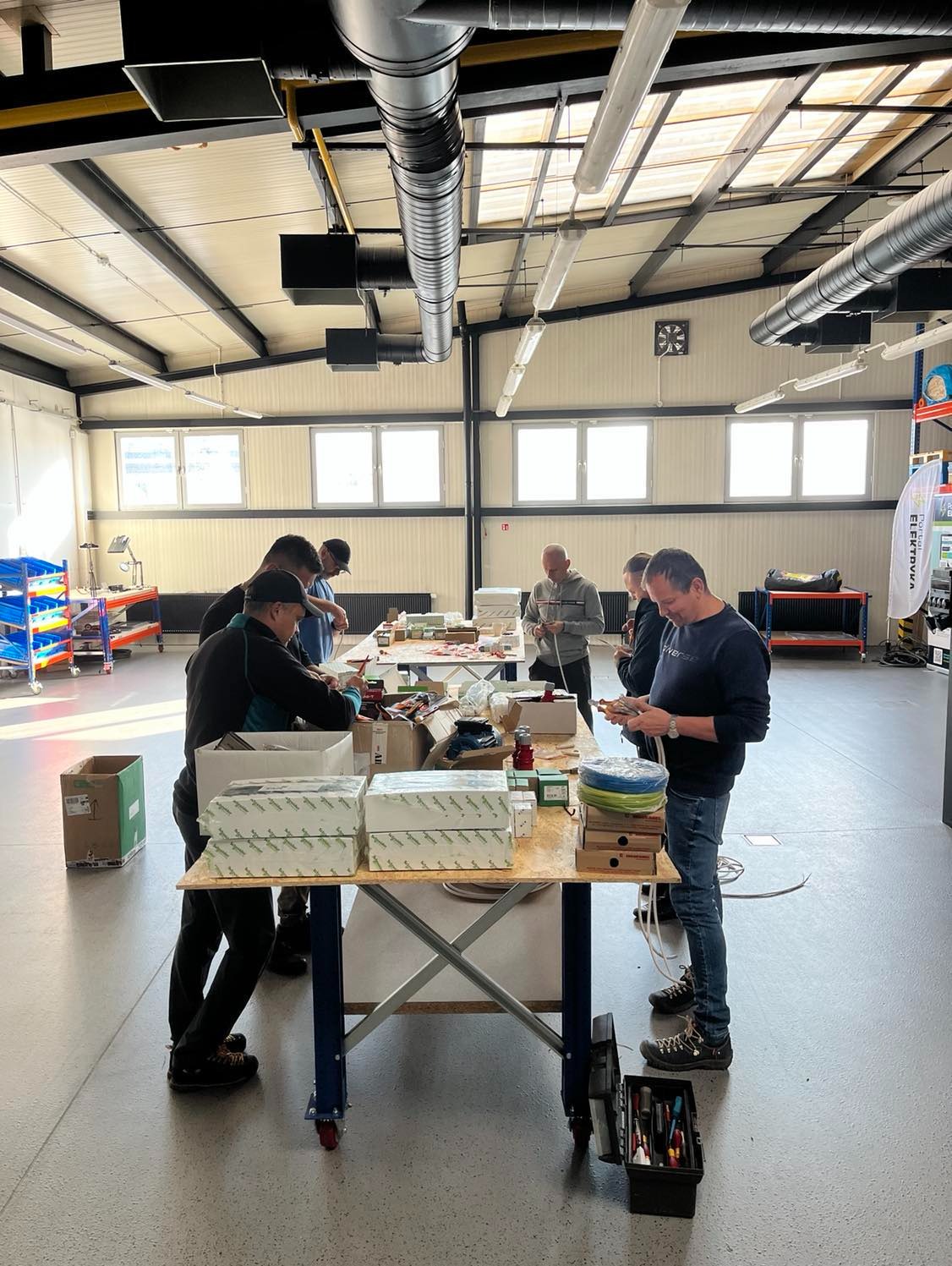 Grupa osób w Mościskach uczestniczy w praktycznym szkoleniu z zakresu instalacji elektrycznych, widoczne stoły z materiałami, narzędzia i skrzynki z osprzętem.