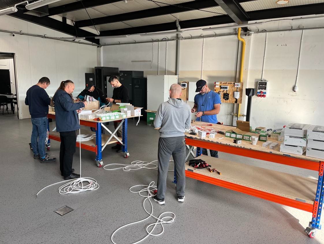 Grupa mężczyzn uczestniczy w praktycznym szkoleniu z zakresu instalacji elektrycznych, pracując przy stołach z elementami instalacji w przestronnym pomieszczeniu.