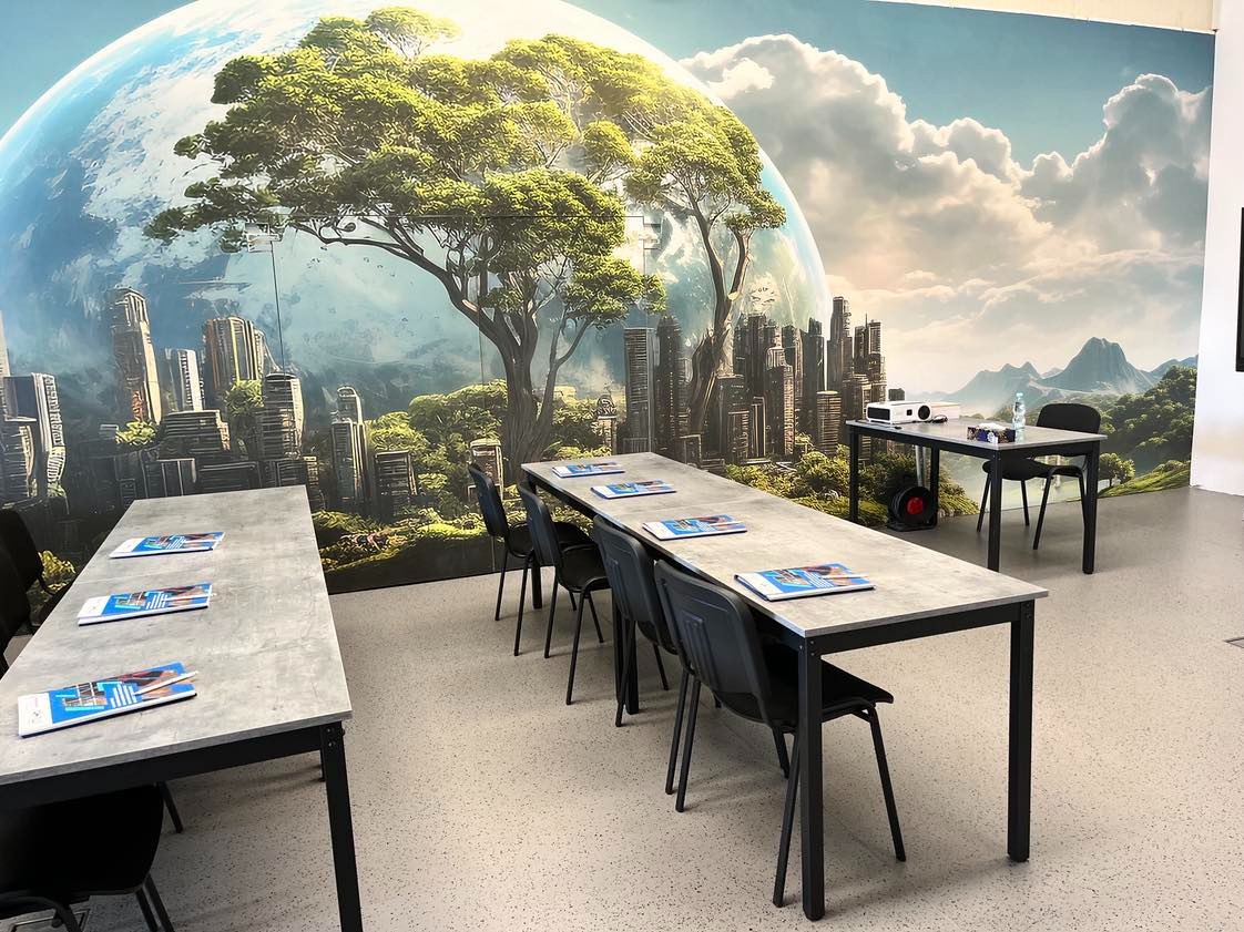 Sala szkoleniowa z długimi stołami, krzesłami, materiałami informacyjnymi i projektorem na tle fototapety przedstawiającej futurystyczny krajobraz z drzewami i drapaczami chmur.