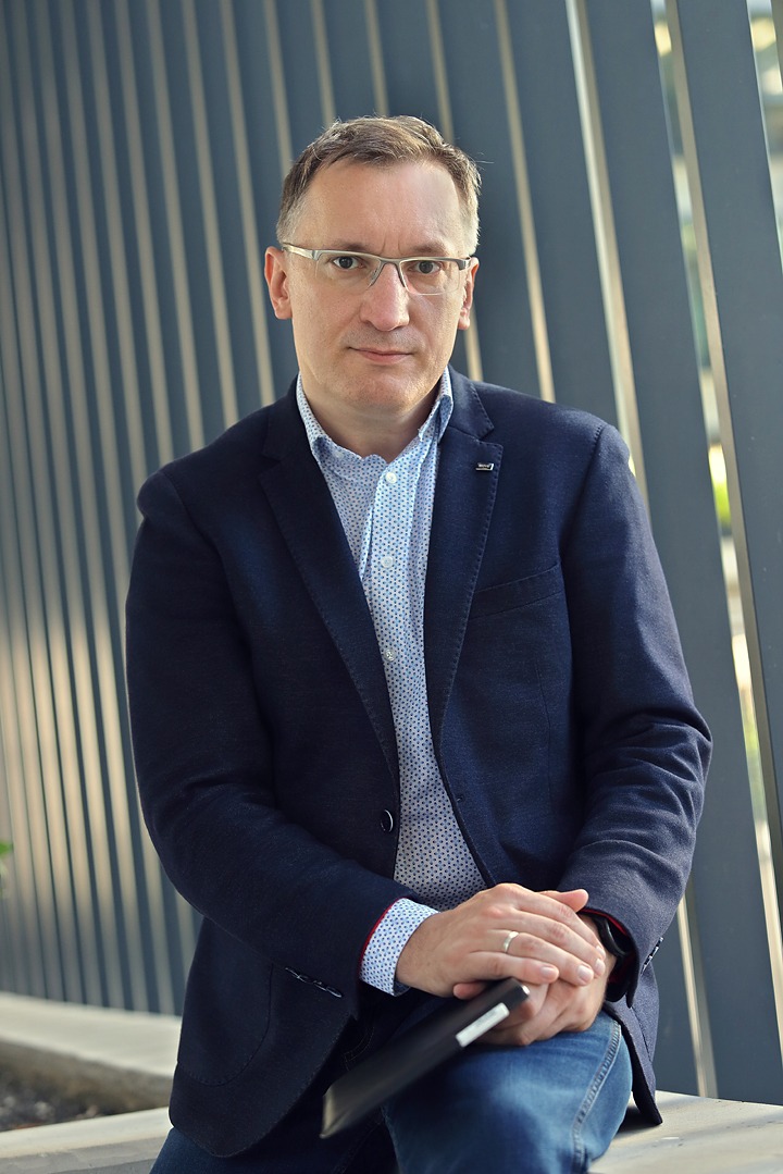 Portret mężczyzny w okularach, ubrany w niebieską marynarkę i wzorzystą koszulę, trzymającego w dłoniach tablet, na tle nowoczesnej konstrukcji z pionowych, szarych elementów.