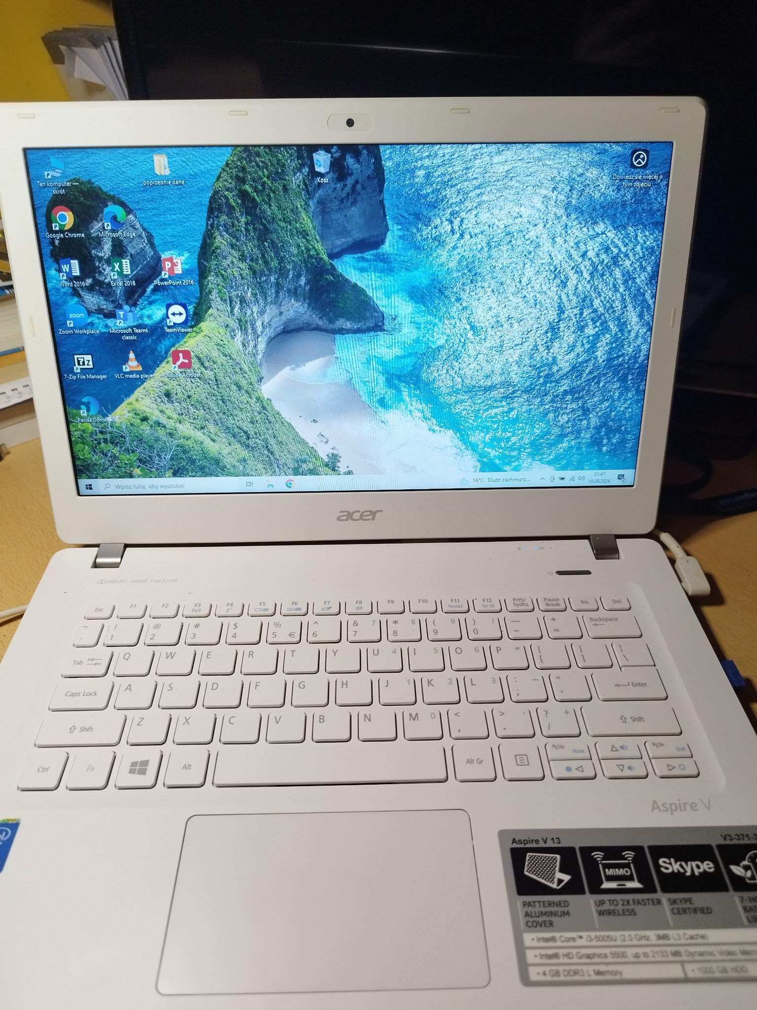 Biały laptop Acer Aspire V z widocznym pulpitem z ikonami programów i tapetą przedstawiającą tropikalną plażę. Widoczne naklejki z informacjami o specyfikacji i certyfikacie Skype.