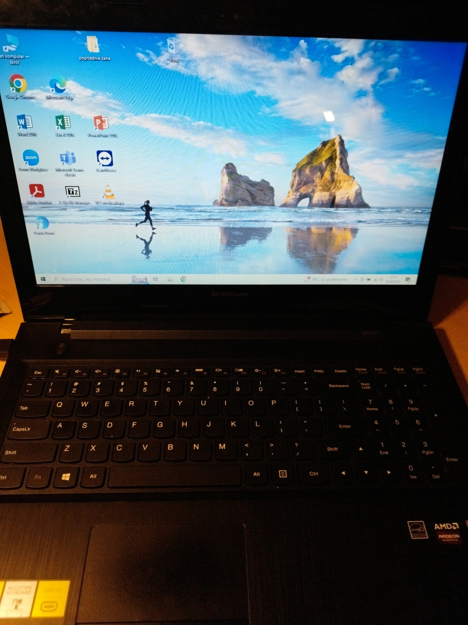 Laptop Lenovo z otwartym ekranem wyświetlającym pulpit z ikonami aplikacji biurowych i graficznych oraz tapetą przedstawiającą osobę biegnącą po plaży z formacjami skalnymi w tle.