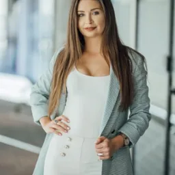 Portret kobiety w jasnym biurze, ubrana w biały top, spodnie z ozdobnymi guzikami i szaro-białą marynarkę w kratę, z zegarkiem i pierścionkiem, patrząca w kamerę.