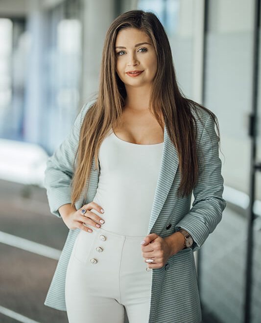 Portret kobiety w jasnym biurze, ubrana w biały top, spodnie z ozdobnymi guzikami i szaro-białą marynarkę w kratę, z zegarkiem i pierścionkiem, patrząca w kamerę.