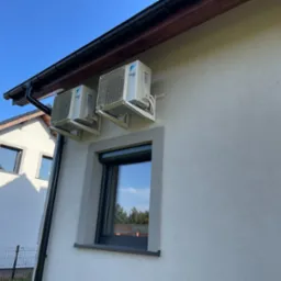 Dwa klimatyzatory Daikin zamontowane na szarej ścianie domu jednorodzinnego, widoczne okno z roletą i fragment sąsiedniego budynku pod błękitnym niebem.