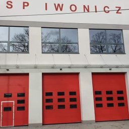 TRENDHOMES Okna i Drzwi - Fasada budynku OSP Iwonicz z trzema czerwonymi bramami garażowymi segmentowymi i oknami z refleksami drzew. Widok z zewnątrz, w dzień.