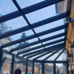 TRENDHOMES Okna i Drzwi - Szklana konstrukcja z czarnymi profilami tworząca zadaszenie tarasu, widok z dołu na błękitne niebo i zieleń drzew. Elementy architektoniczne budynku w tle.
