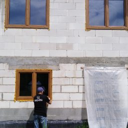TRENDHOMES Okna i Drzwi - Montaż okna z brązową ramą w surowym budynku z betonu komórkowego. Widoczna osoba w trakcie uszczelniania okna pianką montażową. Na ścianie zamontowane 3 okna.