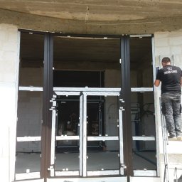 TRENDHOMES Okna i Drzwi - Montaż brązowych drzwi przesuwnych z białymi listwami ochronnymi w surowym stanie budynku. Pracownicy na drabinie podczas instalacji.