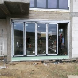 TRENDHOMES Okna i Drzwi - Montaż panoramicznych drzwi przesuwnych w surowym budynku. Widoczni pracownicy podczas instalacji. Duże przeszklenia z białymi ramami.