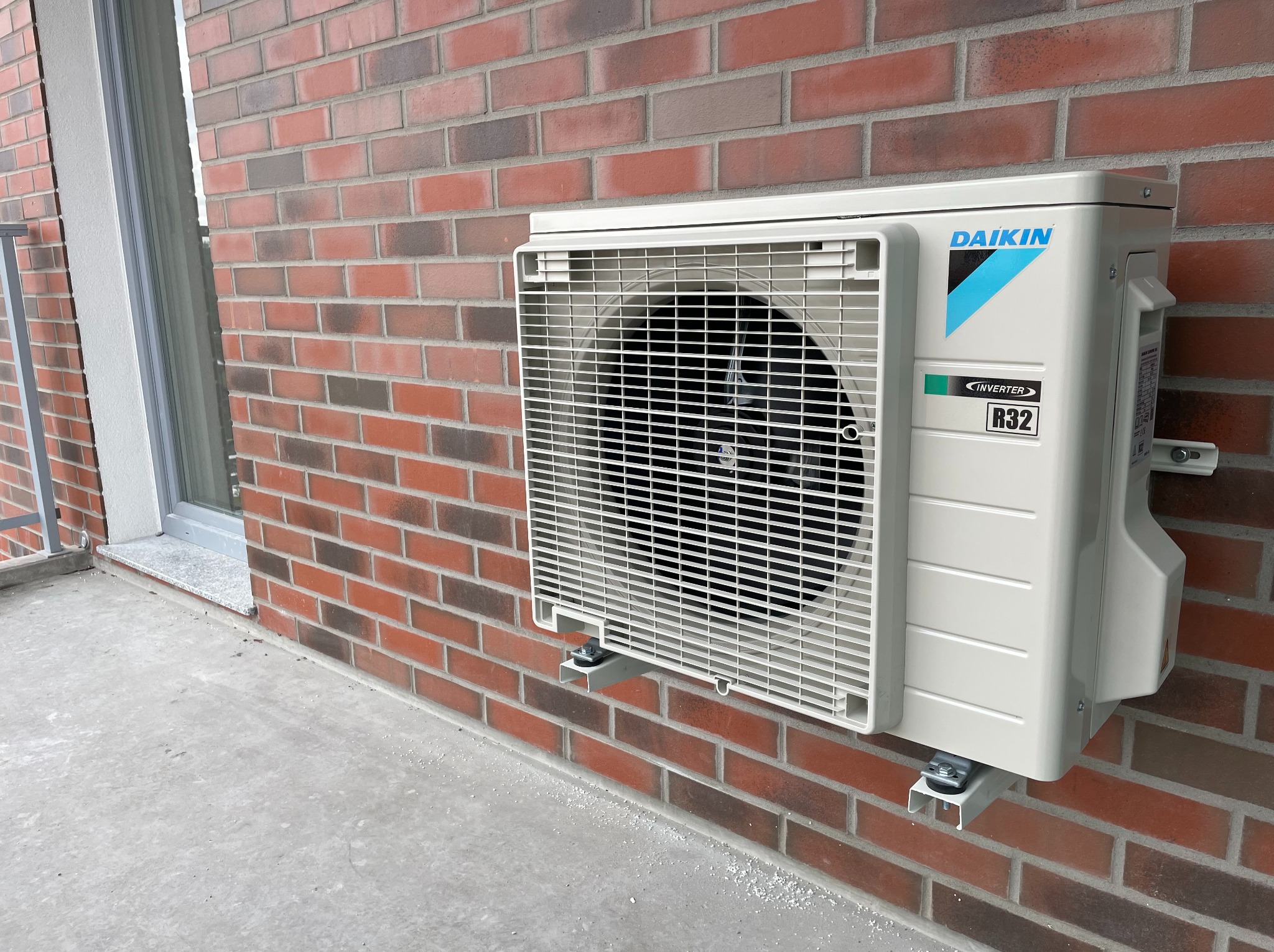 Zewnętrzna jednostka klimatyzacji Daikin zamontowana na ceglanej ścianie budynku, widoczne okno w tle oraz ślady białego pyłu budowlanego na podłożu.