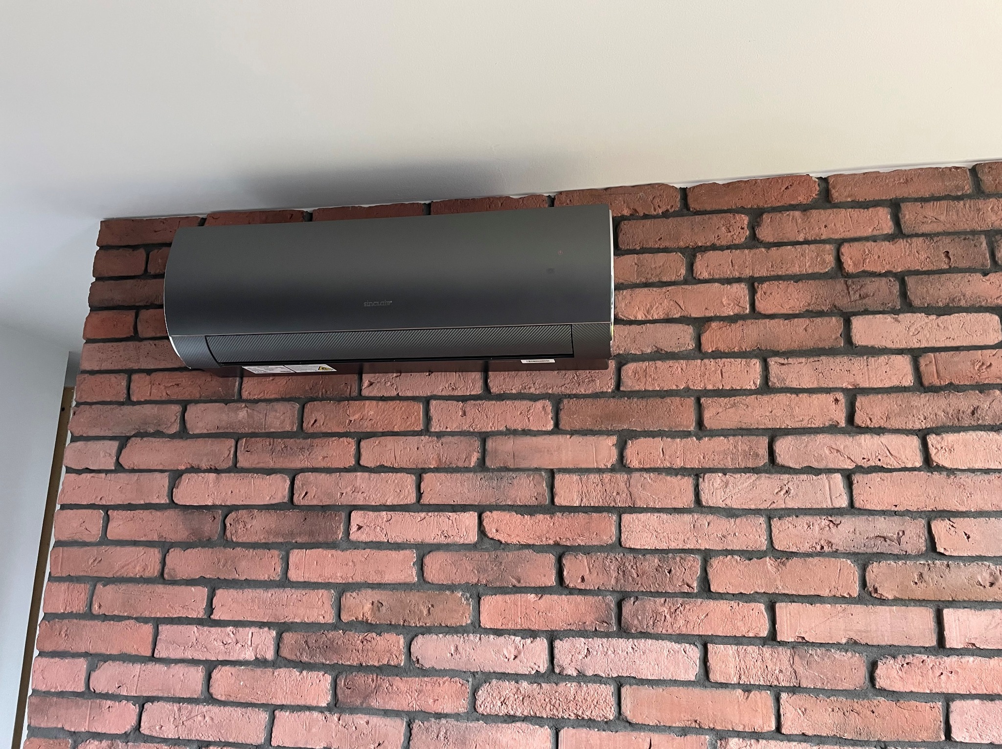 Szara klimatyzacja Sinclair zamontowana na ścianie z czerwonej cegły w pomieszczeniu z białym sufitem.