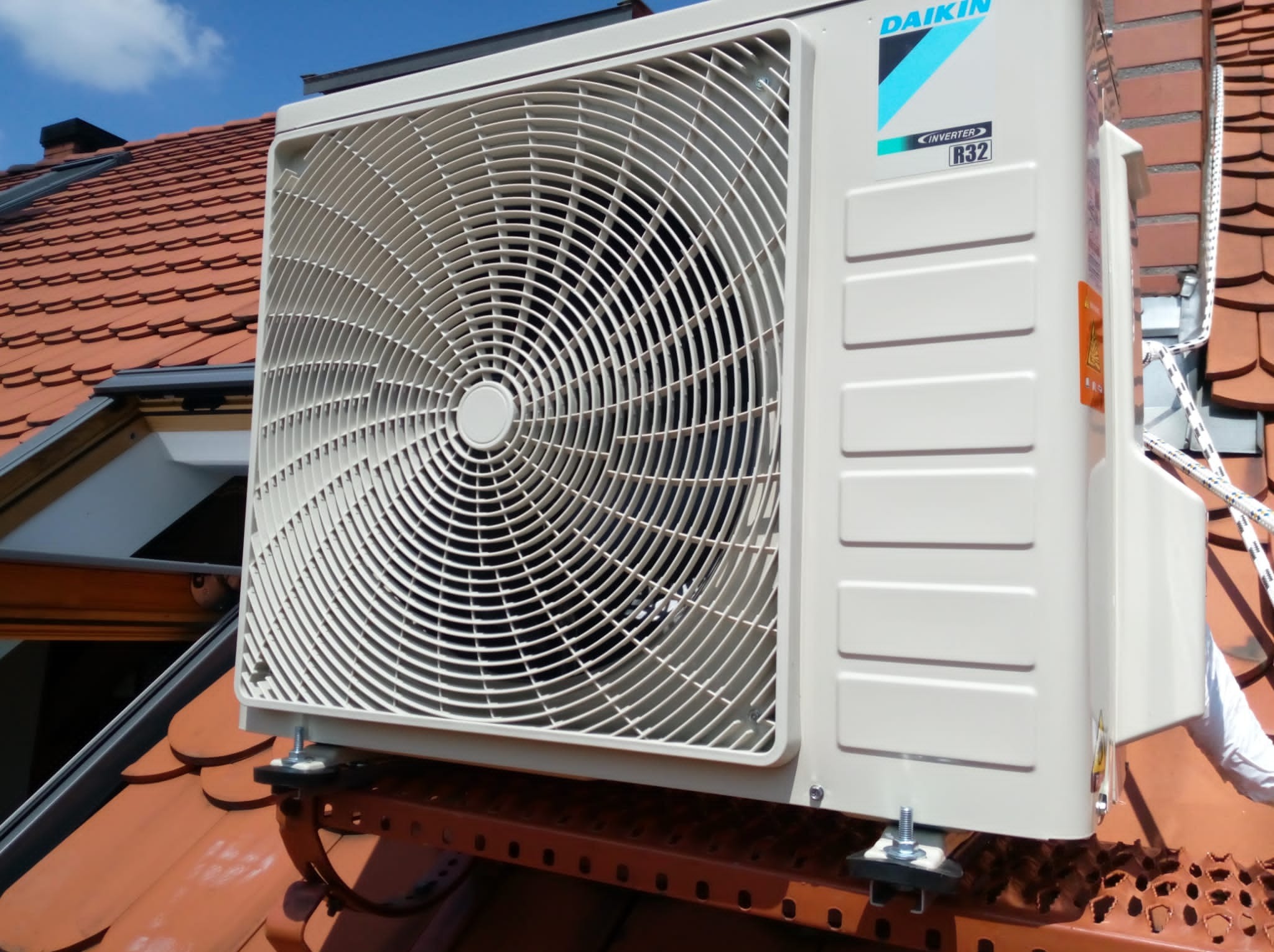 Zewnętrzna jednostka klimatyzacji marki Daikin zamontowana na dachu z czerwonej dachówki, widoczna tabliczka informacyjna R32.