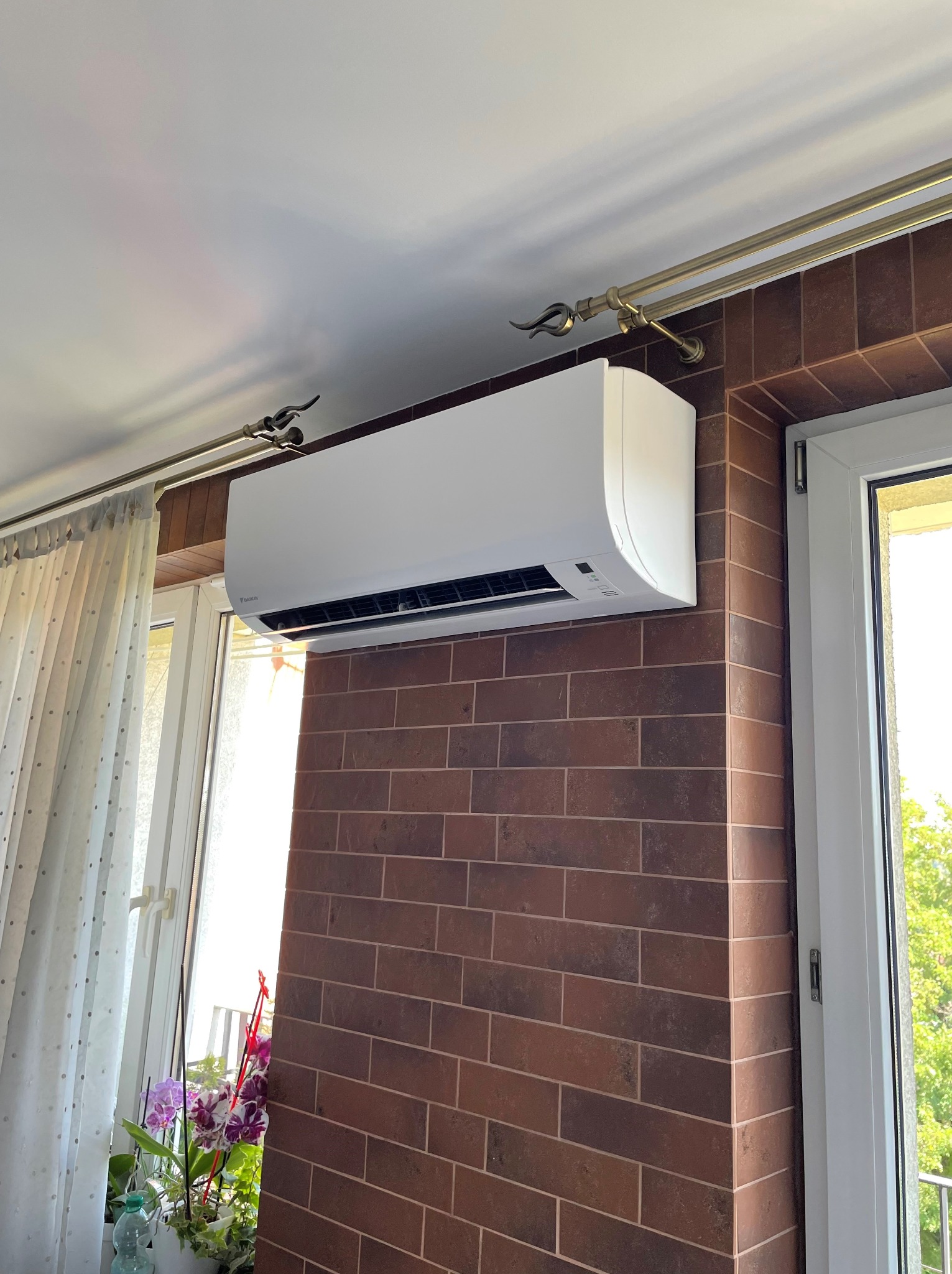 Biała jednostka klimatyzacji zamontowana na ścianie z cegły, widoczna firanka w oknie, dekoracyjny karnisz i kwiaty w doniczce.