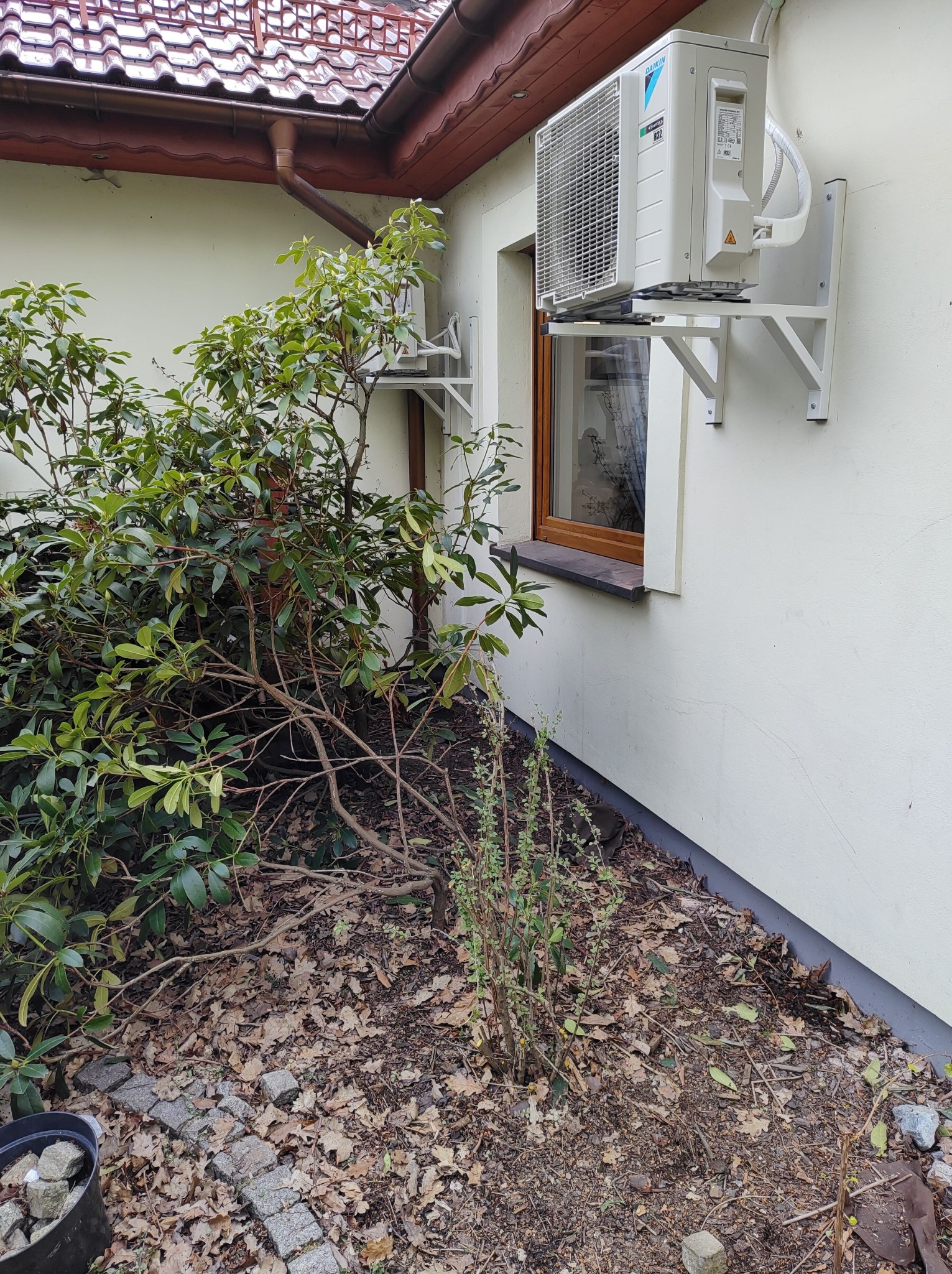 Zewnętrzna jednostka klimatyzacji Daikin zamontowana na białej ścianie budynku, widoczna z poziomu ogrodu z krzewami i opadłymi liśćmi.