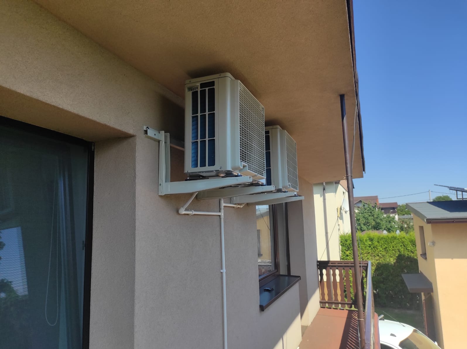 Dwa klimatyzatory zamontowane na elewacji budynku, widoczne rury odprowadzające skropliny, tło z zielenią i błękitnym niebem.