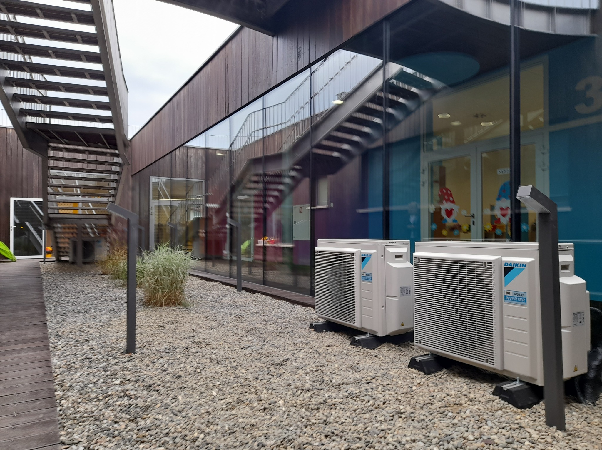 Dwa zewnętrzne agregaty klimatyzacji Daikin Multi Inverter na żwirowym podłożu przed nowoczesnym budynkiem z drewnianą elewacją i szklanymi ścianami odbijającymi metalowe schody.