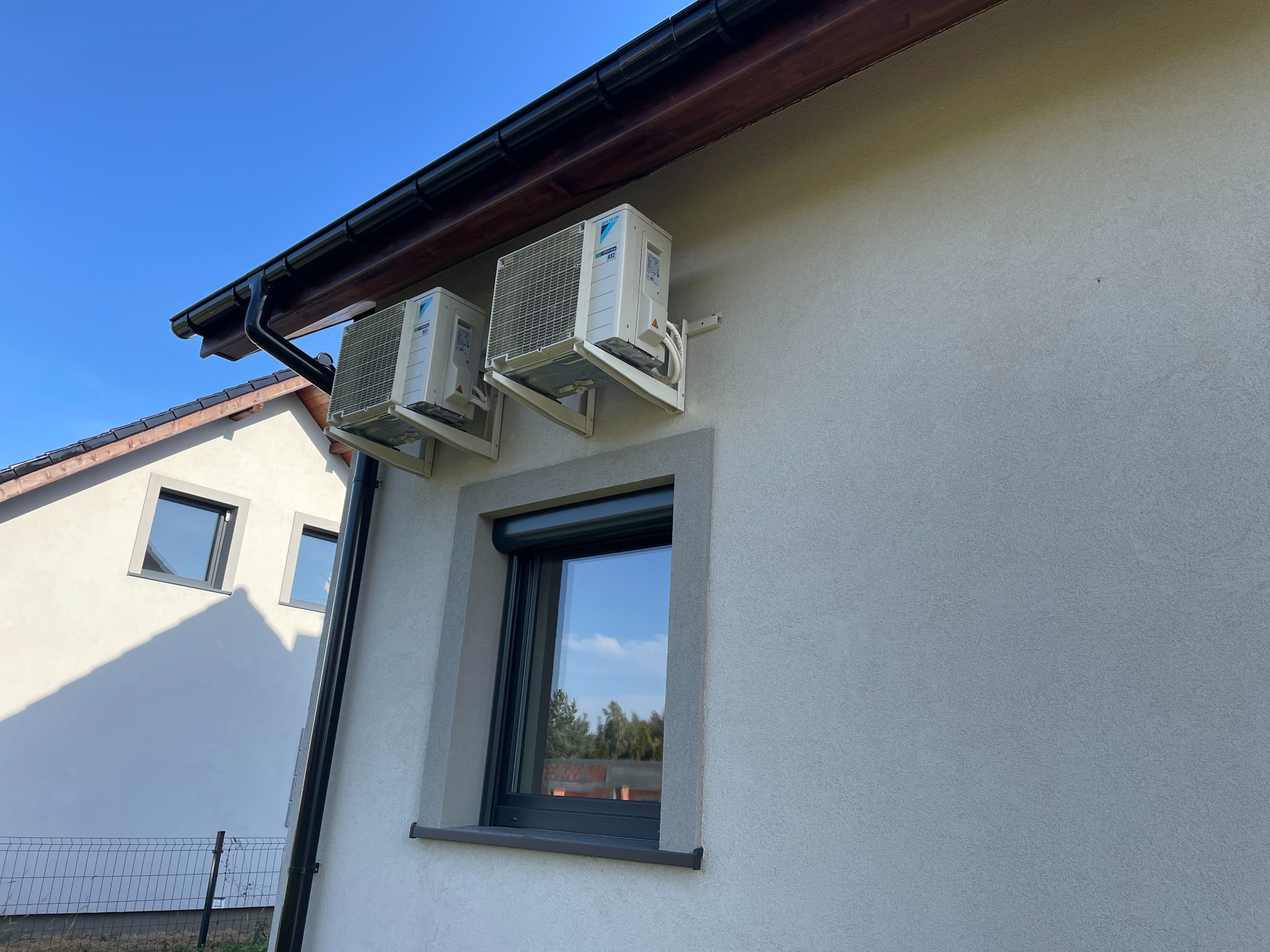 Dwa klimatyzatory Daikin zamontowane na szarej ścianie domu jednorodzinnego, widoczne okno z roletą i fragment sąsiedniego budynku pod błękitnym niebem.