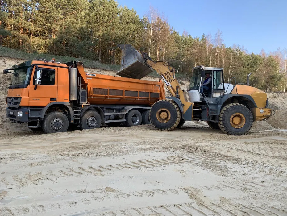 Ładowarka czołowa Liebherr 554 ładuje pomarańczową wywrotkę Mercedes-Benz 4148 na piaszczystym terenie, widoczne ślady opon i las w tle.