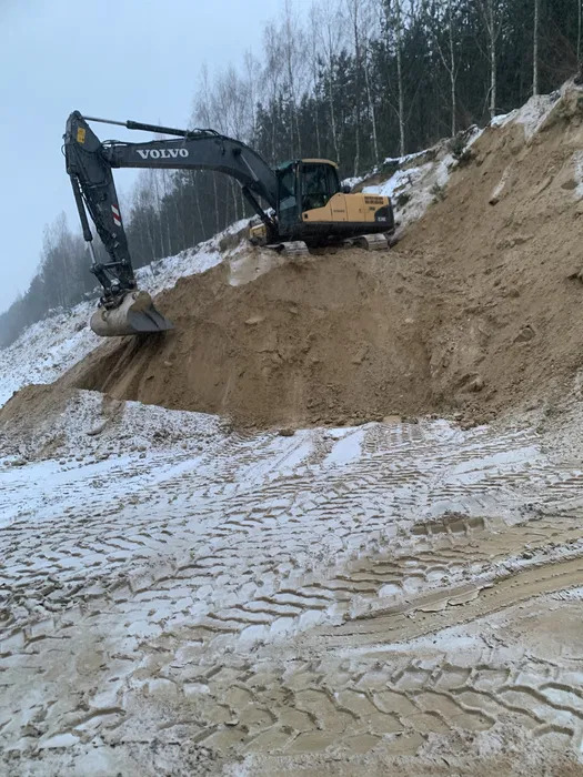 Żółto-czarna koparka Volvo na zboczu ziemi pokrytym śniegiem, ślady opon na śniegu, w tle las i pochmurne niebo.