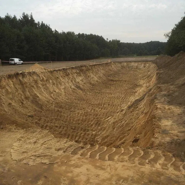 Szeroki wykop w ziemi z widocznymi śladami opon maszyn budowlanych na dnie i skarpach, zabezpieczony taśmą ostrzegawczą, w tle las i biały samochód dostawczy.
