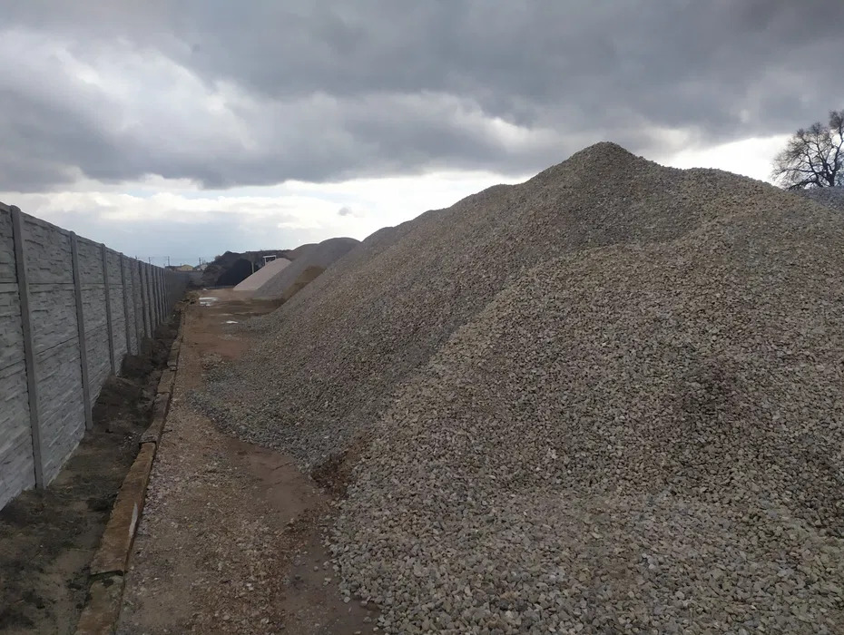 Duża pryzma szarego kruszywa na placu budowy, widoczny betonowy płot i pochmurne niebo.