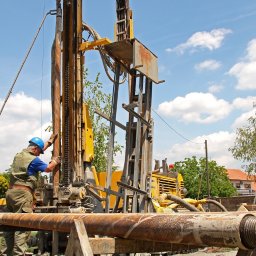 GeoTerm Wrocław - Dwóch pracowników w kaskach obsługuje wiertnicę na tle błękitnego nieba z chmurami; widoczne rury wiertnicze i zabudowania w oddali.