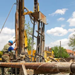 Dwóch pracowników w kaskach obsługuje wiertnicę na tle błękitnego nieba z chmurami; widoczne rury wiertnicze i zabudowania w oddali.