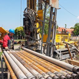 GeoTerm Wrocław - Dwóch pracowników w roboczych strojach obsługuje żółtą wiertnicę do studni głębinowej; na pierwszym planie ułożone rury wiertnicze, w tle domy jednorodzinne i zieleń.