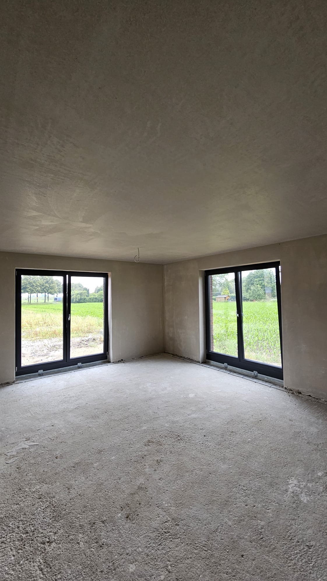 Surowe wnętrze po nałożeniu tynku maszynowego, widoczne dwa okna z widokiem na zewnątrz, surowa podłoga i wystający kabel elektryczny z sufitu.