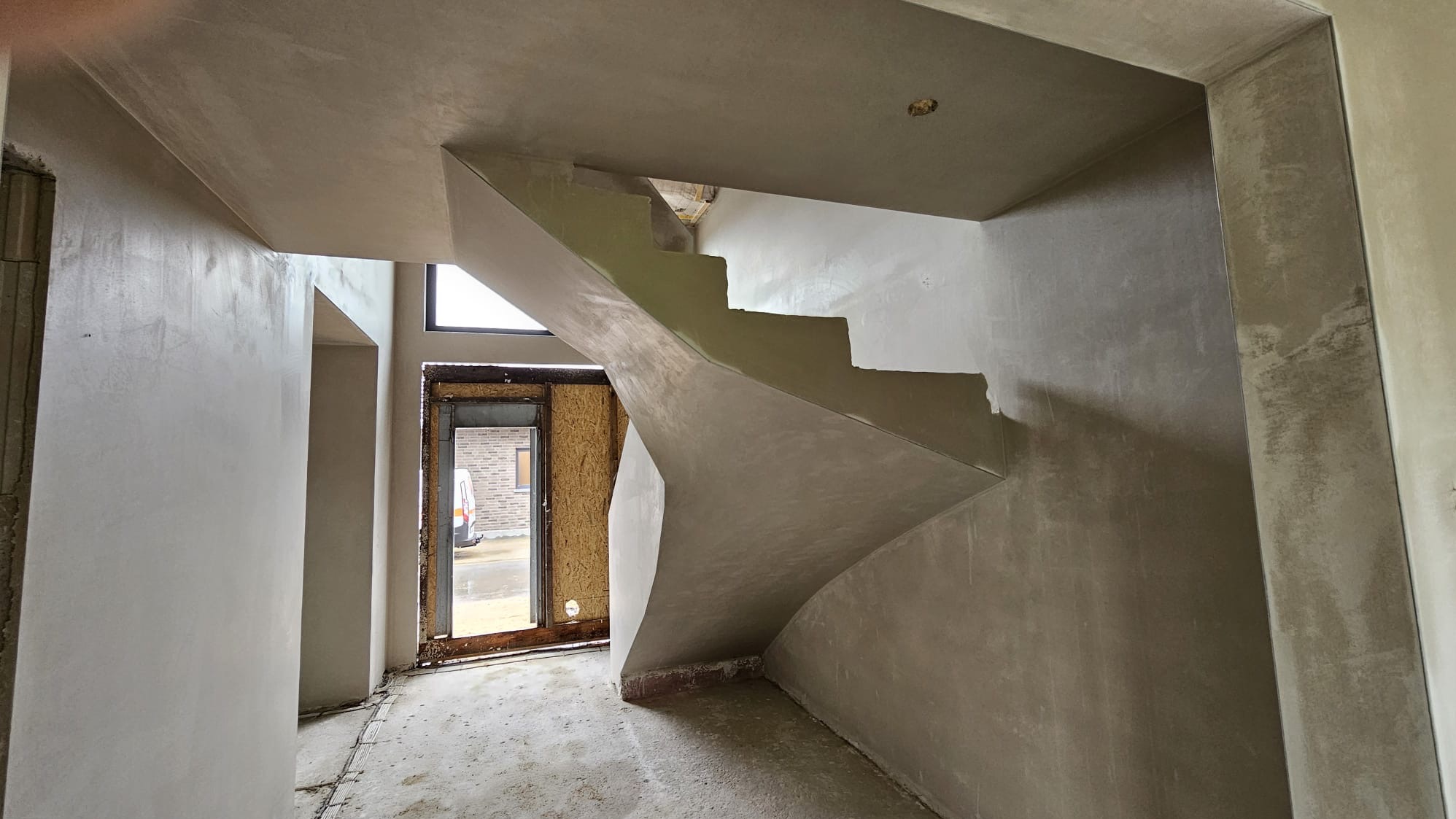 Wnętrze w trakcie prac wykończeniowych: świeżo otynkowane ściany, sufit i schody spiralne, widoczne przez otwarte drzwi fragment ulicy z zaparkowanym busem.