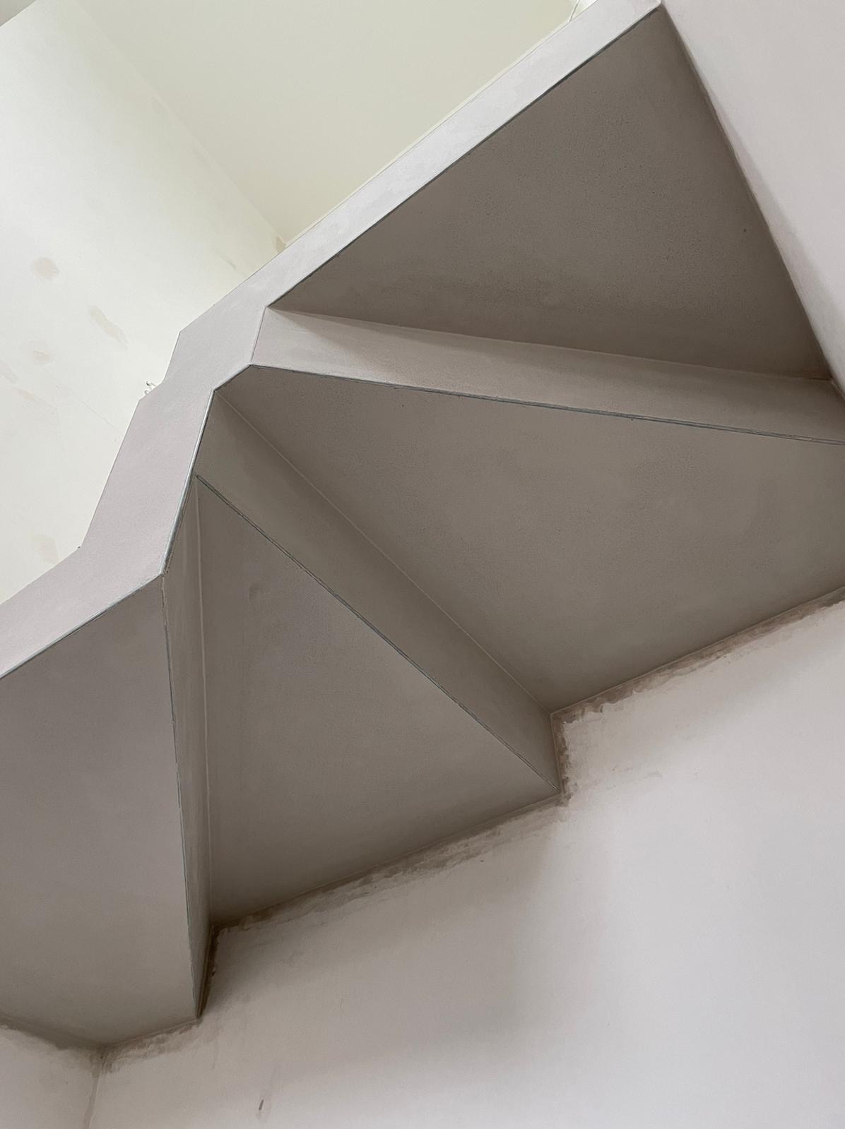 Gładka, szara powierzchnia świeżo nałożonego tynku na skosach sufitu i ścian, widoczne łączenia i niedoskonałości w narożnikach.