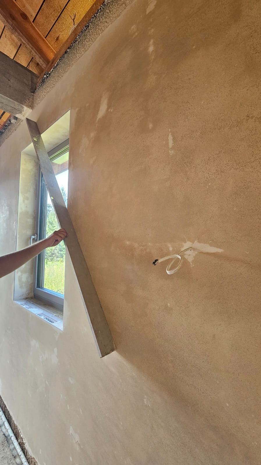 Świeżo nałożona, jasnobrązowa warstwa tynku na ścianie wewnętrznej, wyrównywana długą, szarą łatą przy oknie; widoczne wystające kable elektryczne.