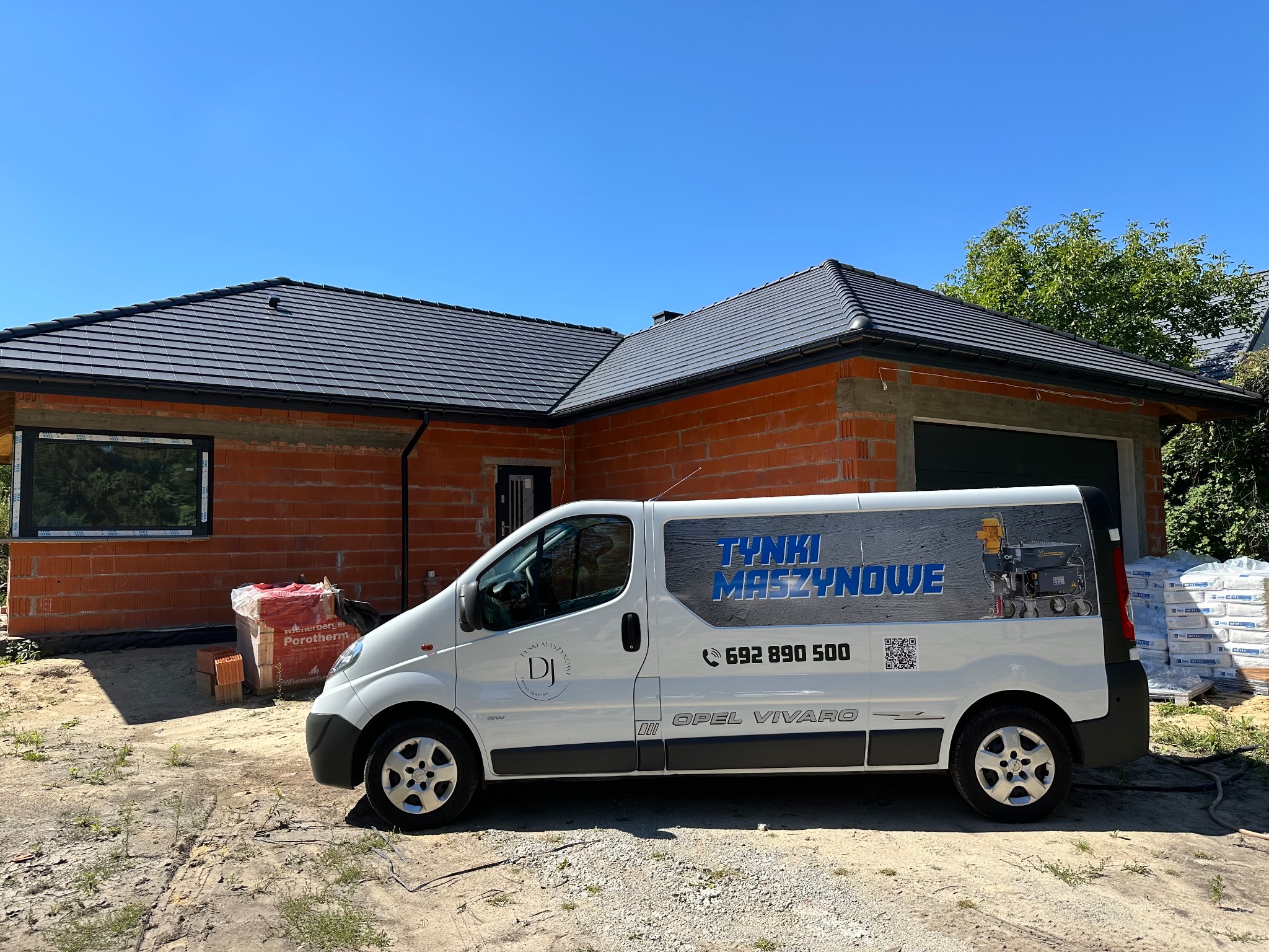 Nowy dom w trakcie budowy, z zaparkowanym białym Oplem Vivaro z reklamą firmy wykonującej tynki maszynowe, obok paleta z bloczkami Porotherm.