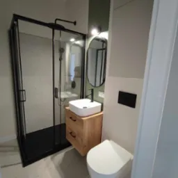 Nowoczesna łazienka z czarną kabiną prysznicową, umywalką na drewnianej szafce z szufladami i owalnym lustrem z czarną ramą, na tle zielonej ściany.