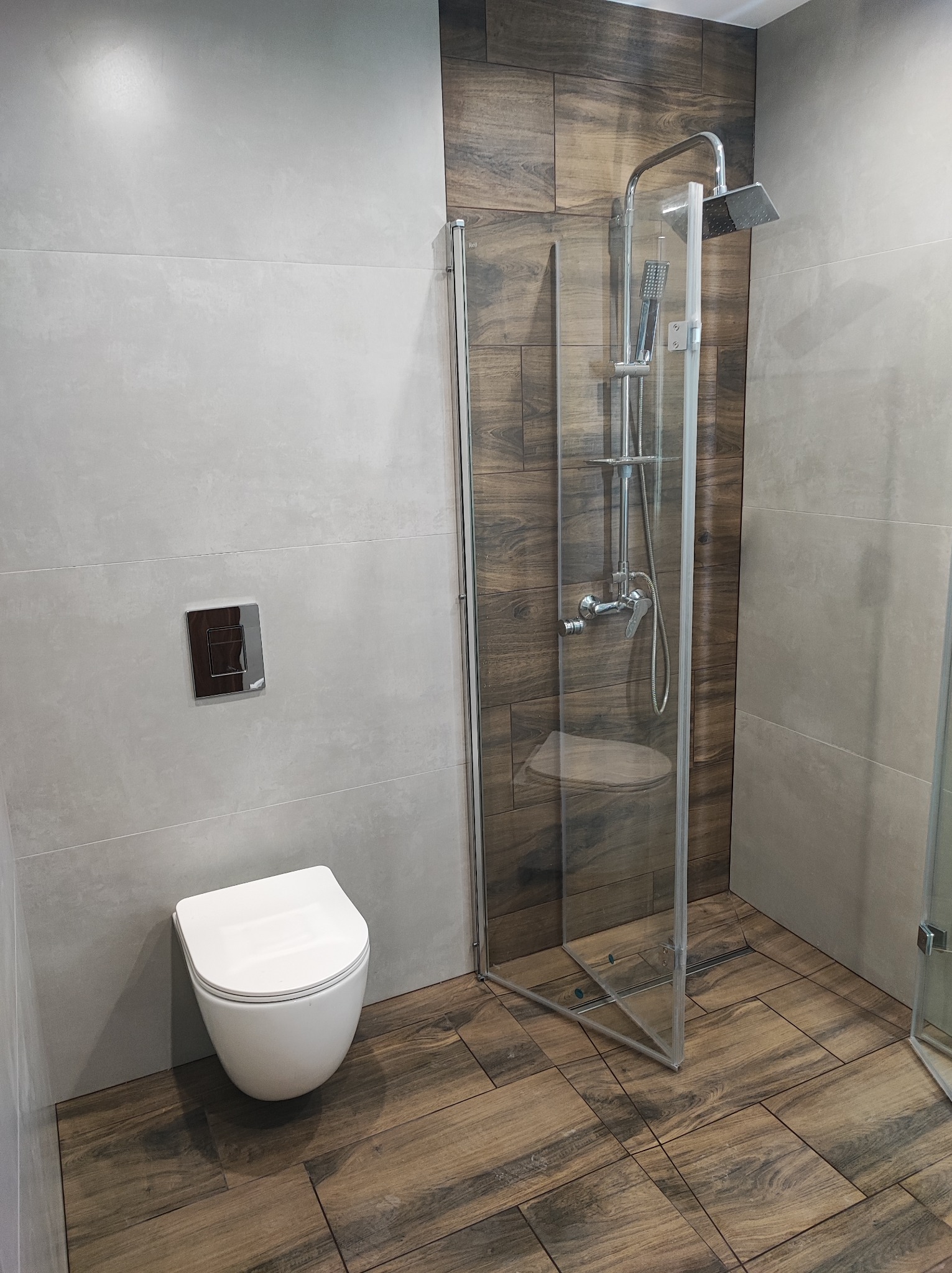 Nowoczesna łazienka z kabiną prysznicową typu walk-in, wiszącą toaletą i płytkami imitującymi drewno na podłodze oraz ścianie w strefie prysznica, kontrastującymi z szarymi płytkami na pozostałych...